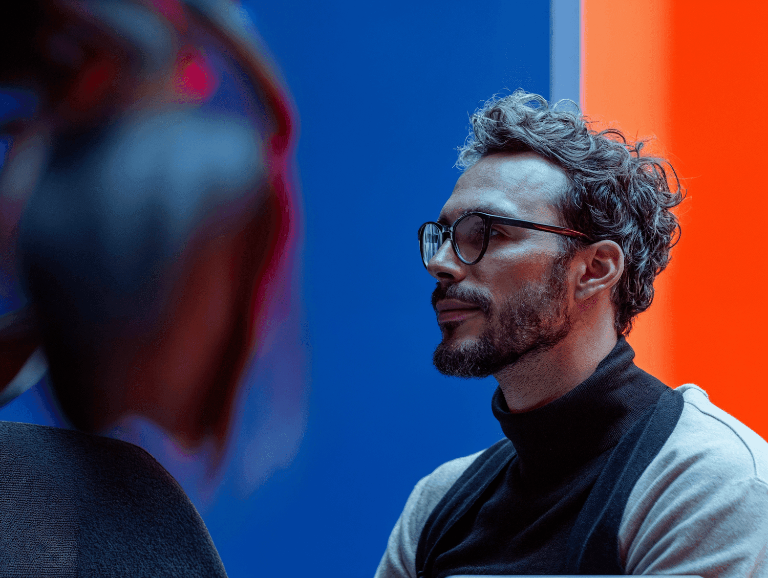 Man with glasses in a turtleneck against blue and orange walls